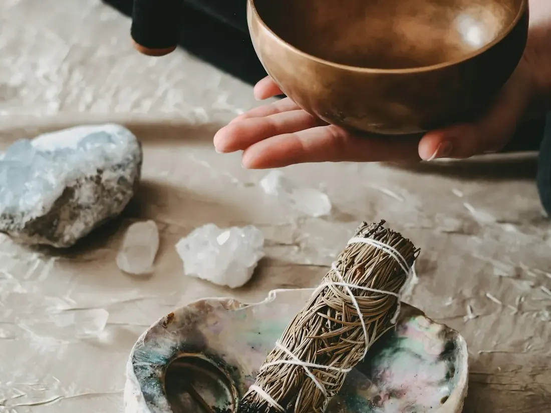 Hand held over a singing bowl beside sage and stones